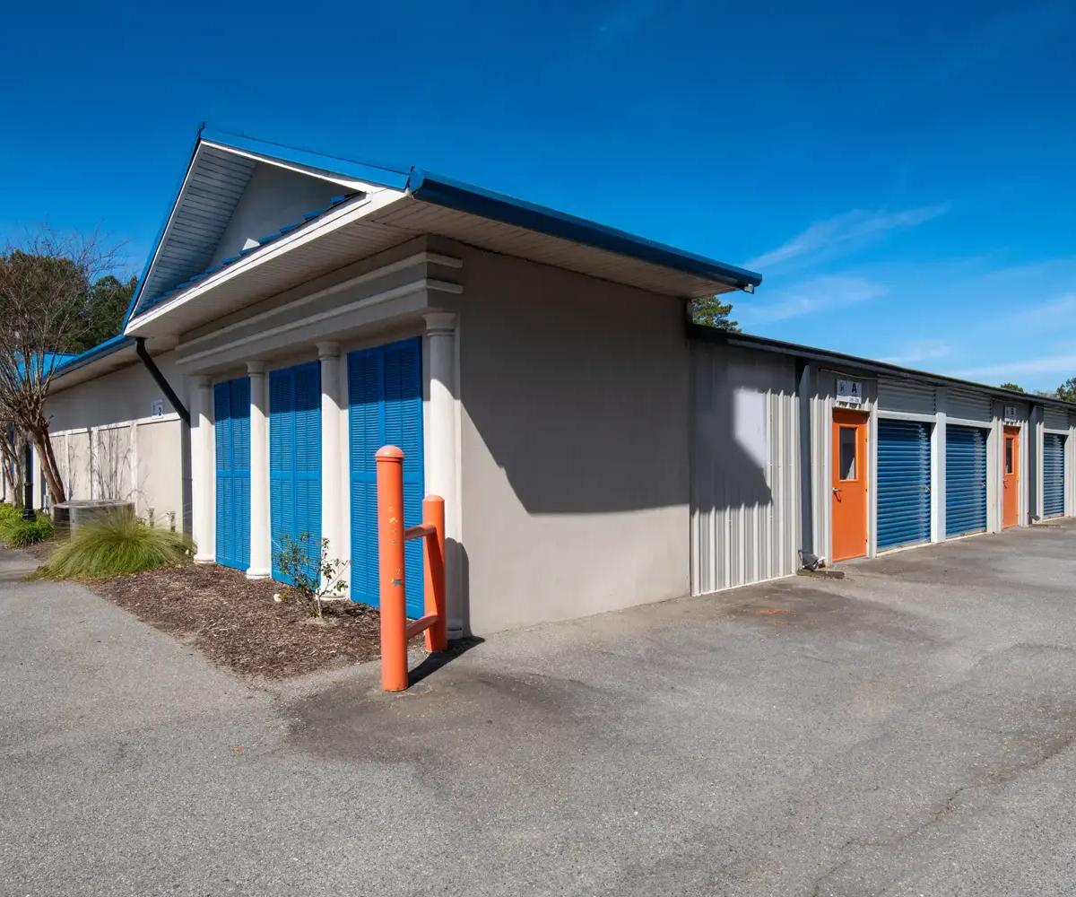 Exterior view of storage unit building with blue roll-up doors and orange entry doors under a clear sky