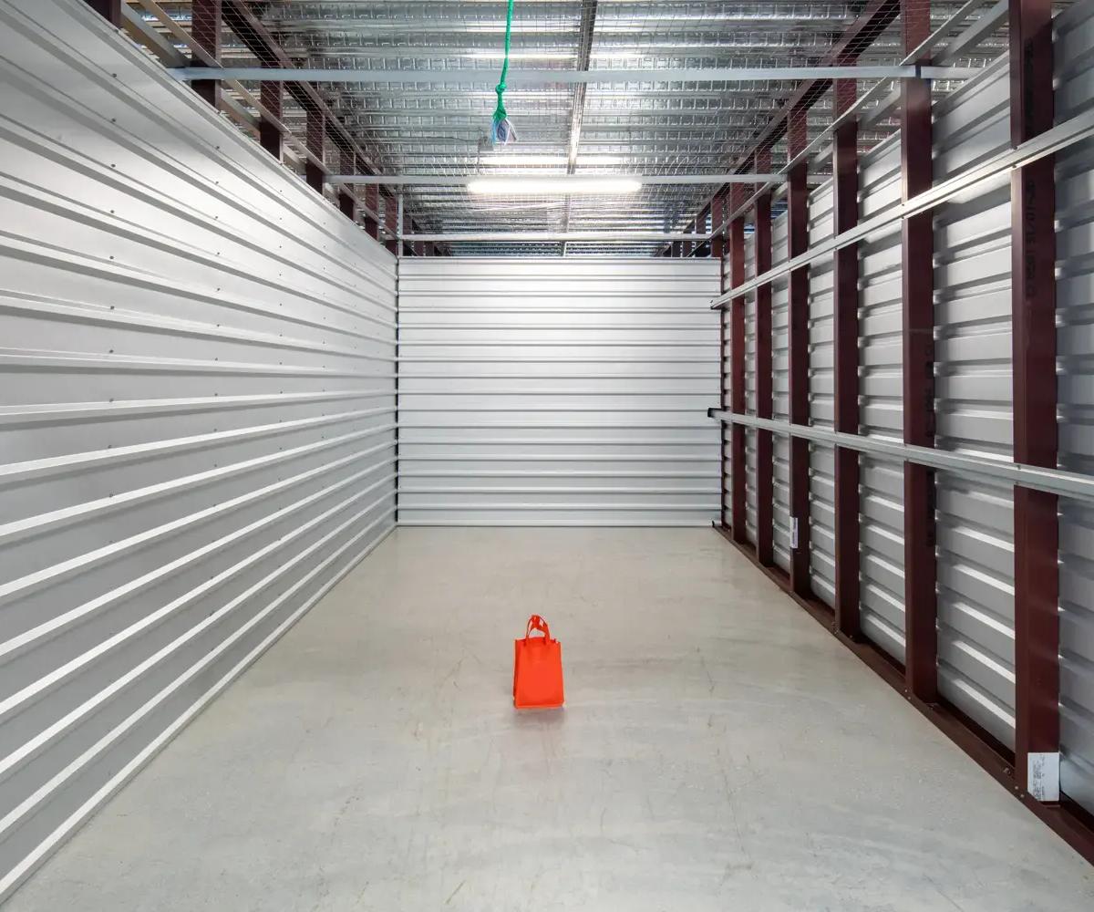 Empty self-storage unit with metal walls, a concrete floor, and one small orange bag in the center