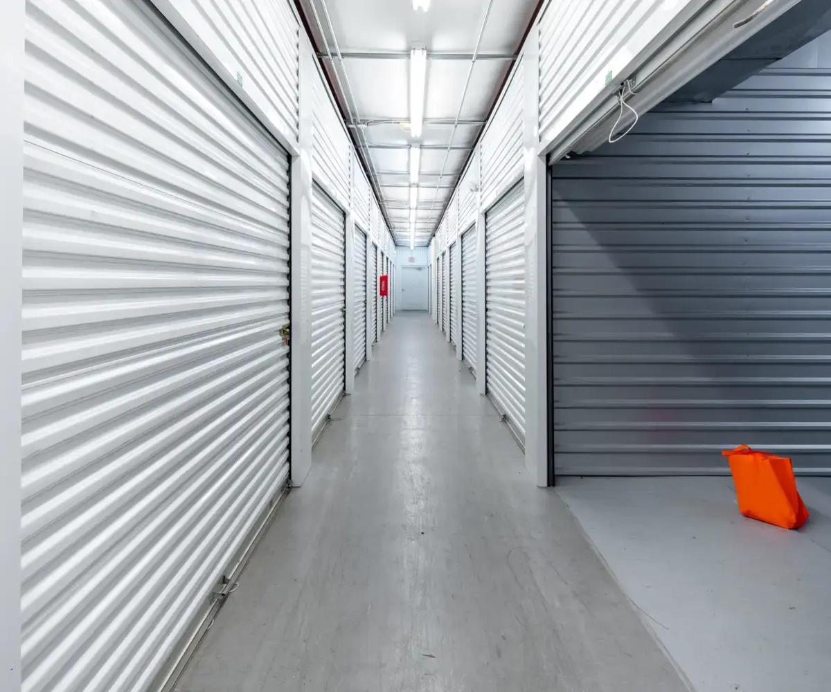 An indoor storage hallway with white roll-up unit doors and an orange bag on the floor