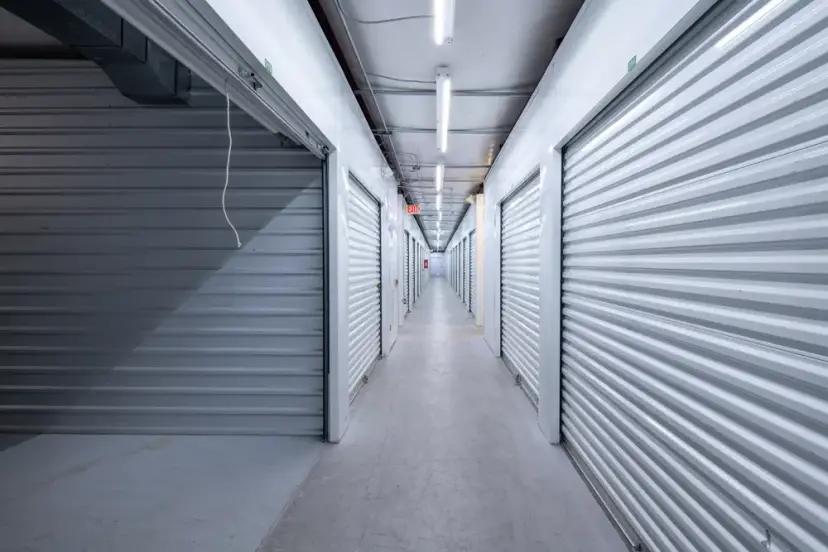 Brightly lit interior hallway with white corrugated metal roll-up storage unit doors and overhead fluorescent lighting