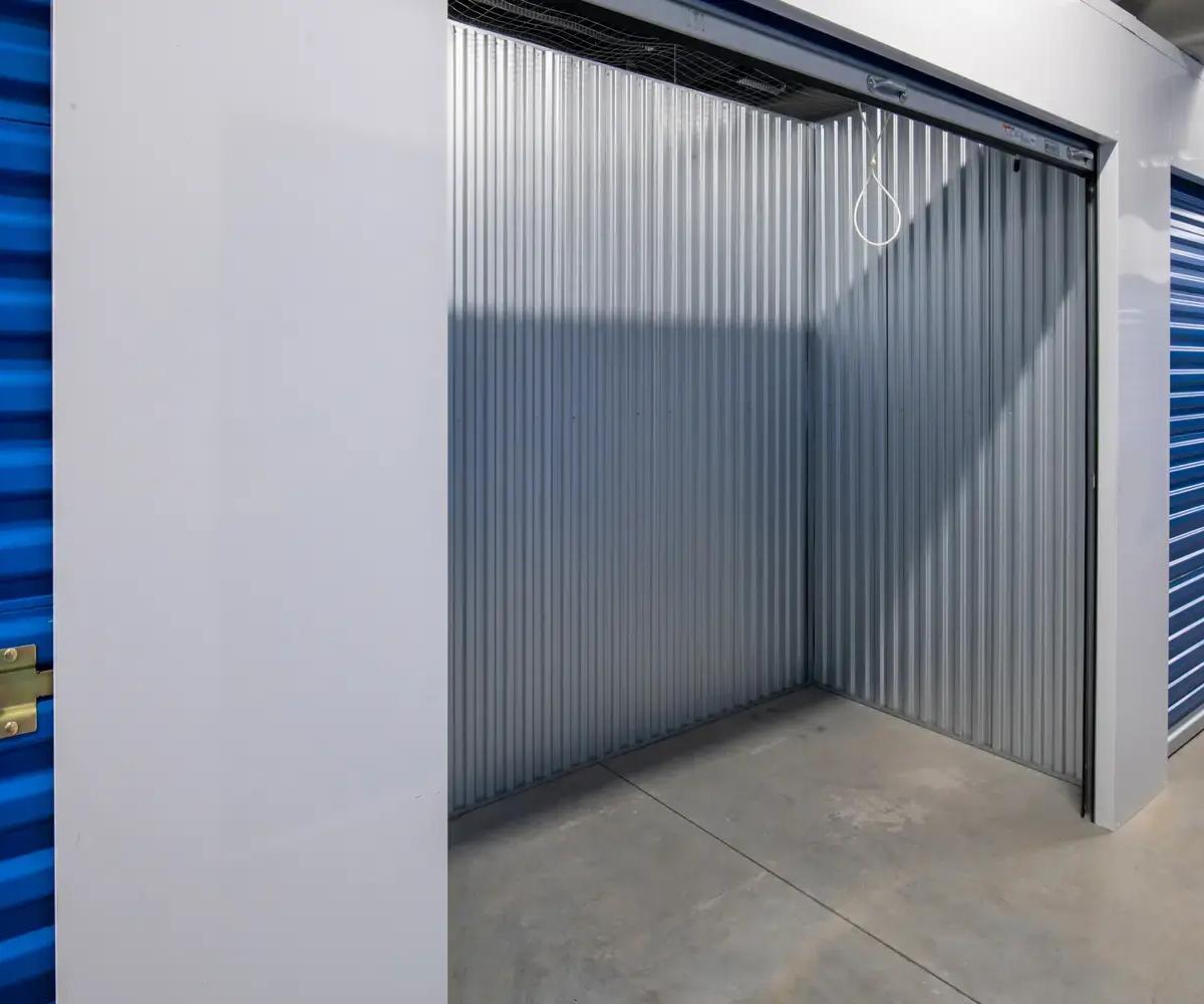 An empty storage unit with corrugated metal walls and a gray concrete floor between blue doors