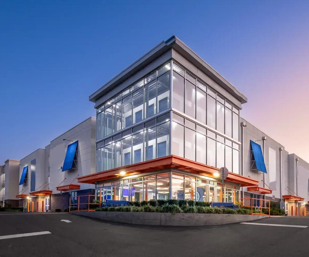 Multi-story self-storage building featuring glass corner walls at dusk with illuminated indoor units and blue awnings