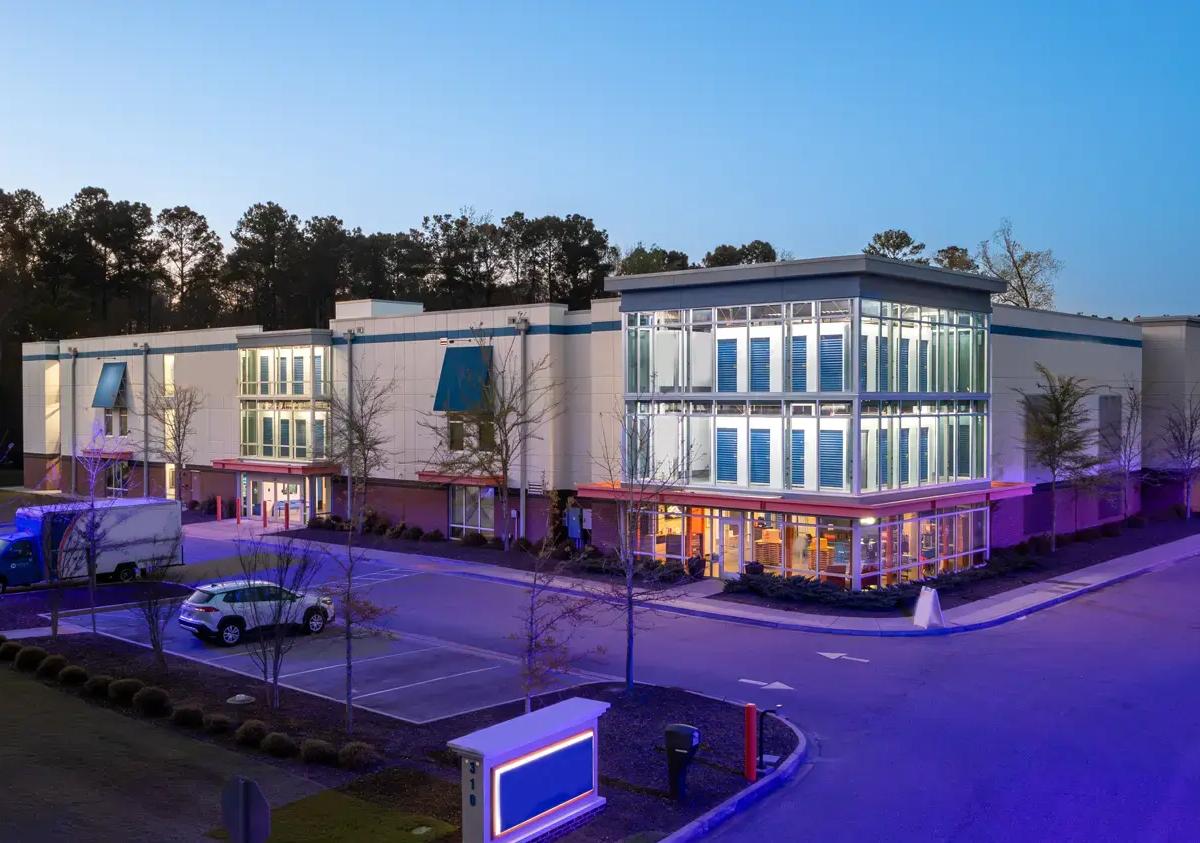 Modern multi-story self-storage facility with glass windows showing interior blue unit doors at dusk