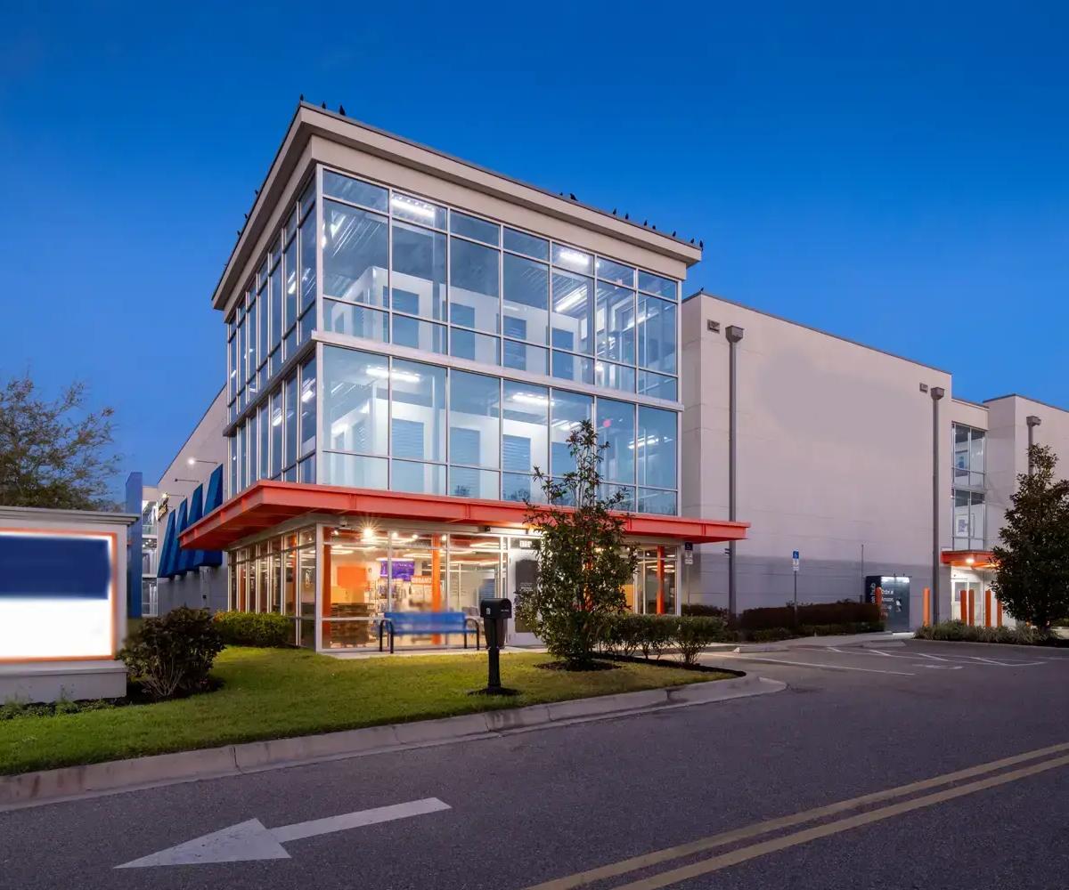 Multi-story storage facility exterior with a glass-front office and orange overhang at twilight