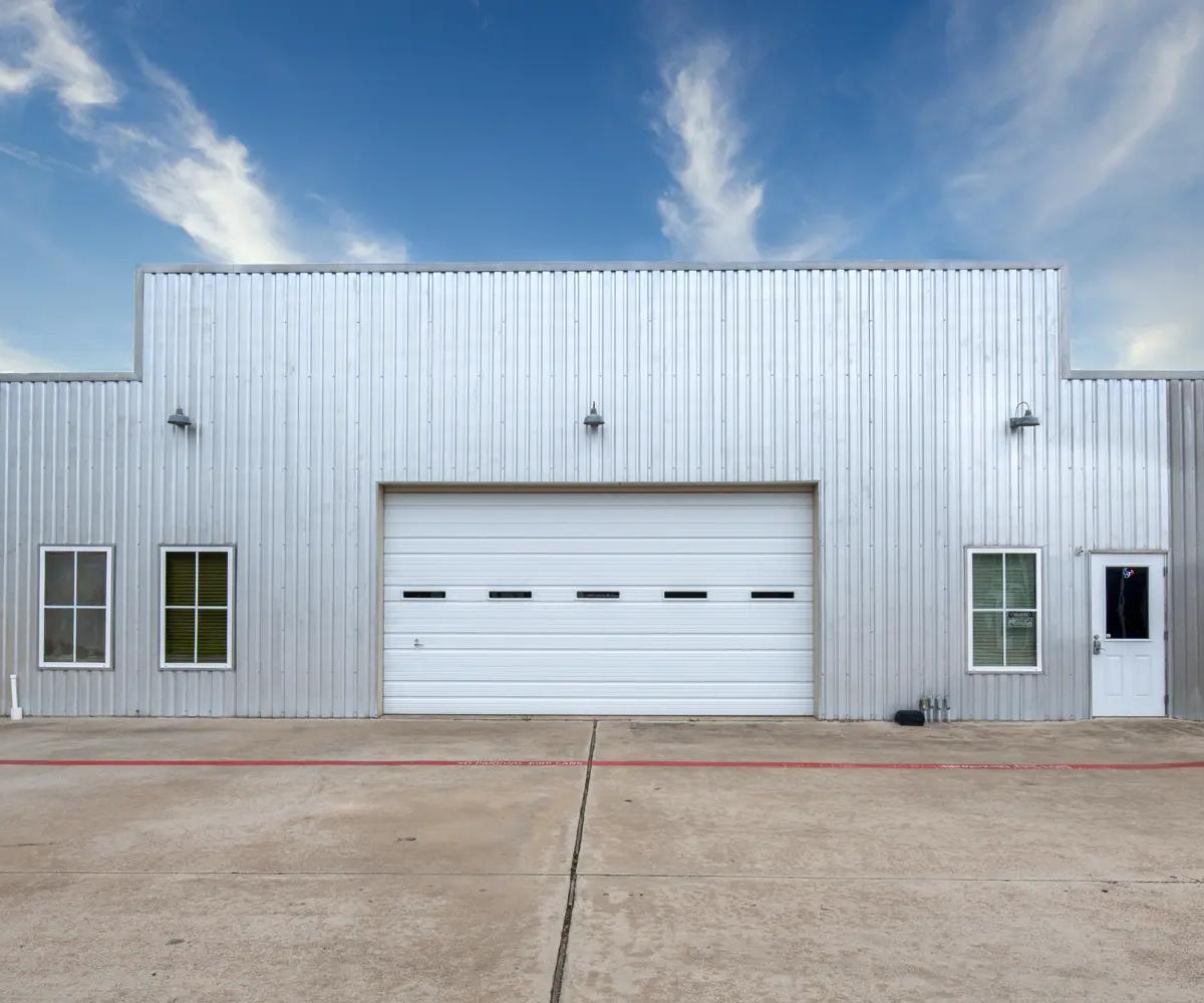 Corrugated metal storage building exterior with a white roll-up door, side windows, and a paved driveway