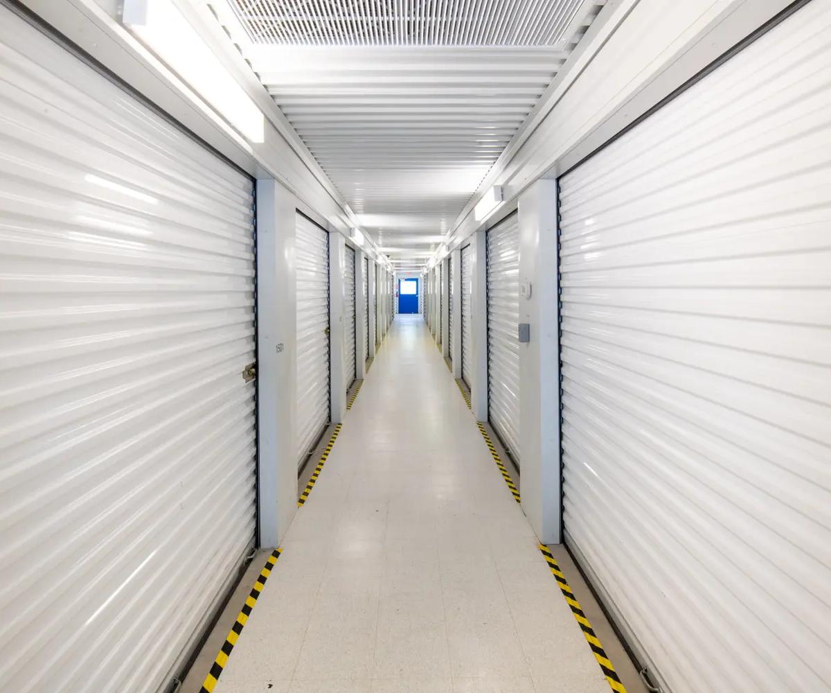 Brightly lit indoor self-storage hallway with white roll-up doors and yellow hazard floor tape leading toward a blue door