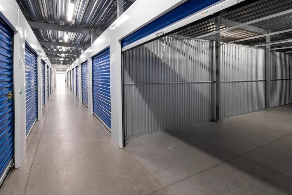 Indoor storage hallway featuring rows of blue roll-up doors and a large open unit with corrugated walls